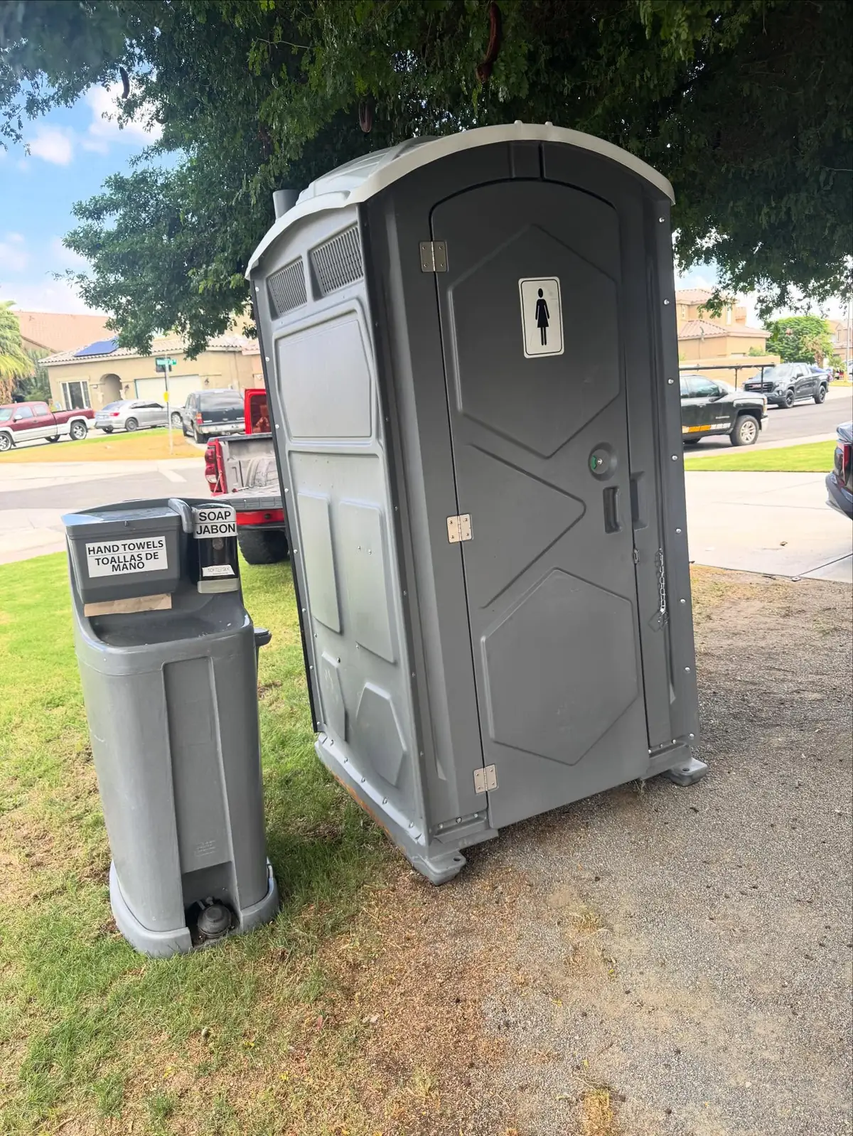 Grey porta potty next to a foot-pump hand-wash station on grass